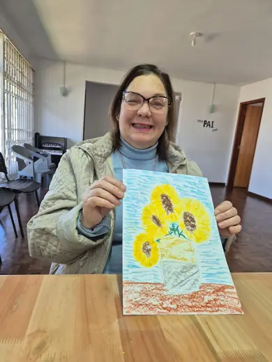 Senhora sorrindo, segurando o desenho de girassóis que acabou de pintar na oficina de artes