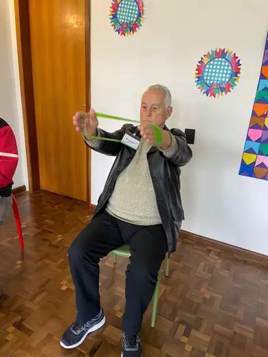 Senhor sentado, fazendo exercício de braço com faixa elástica na aula de atividade física