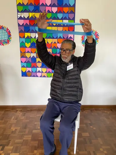 Senhor sentado erguendo uma faixa elástica acima da cabeça, diante do painel de corações coloridos