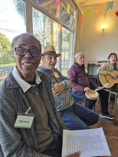 Senhor de óculos sorrindo com a letra da música na mão, e ao fundo o grupo com violão e pandeiro
