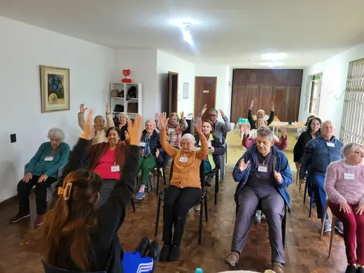 Roda de idosos numa sala ampla, com os braços levantados durante a atividade em grupo