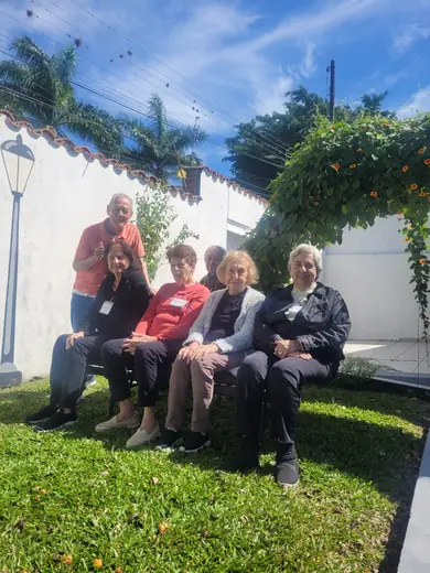 Grupo de idosos e equipe sentados num banco do jardim da casa, em dia de sol
