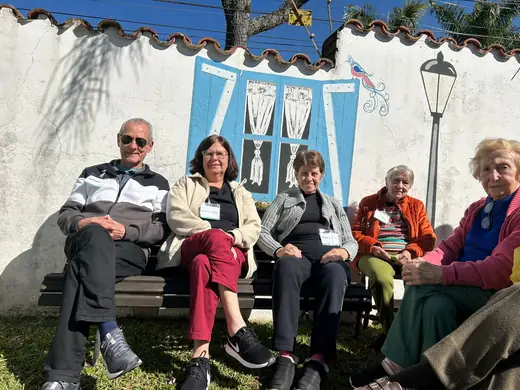 Cinco idosos sentados lado a lado num banco, junto ao muro pintado com uma janela azul