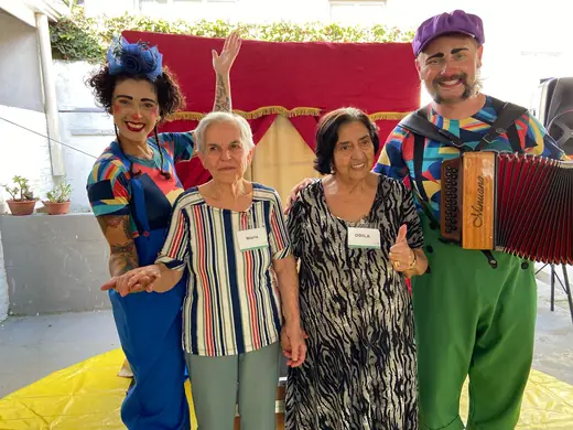 Duas senhoras ao lado de dois artistas de circo, um deles com sanfona, em festa temática no pátio