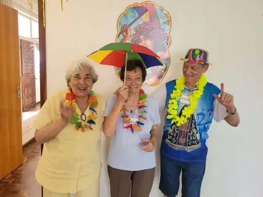Três senhoras animadas no baile, com sombrinha colorida, colar de flores e chapéu