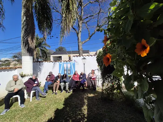 Roda de idosos sentados no jardim ensolarado, com uma trepadeira de flores laranja em primeiro plano