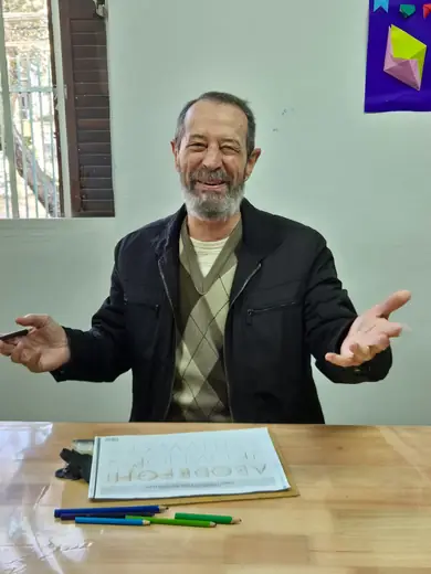 Senhor de barba sorrindo de braços abertos à mesa, com a folha da atividade à frente